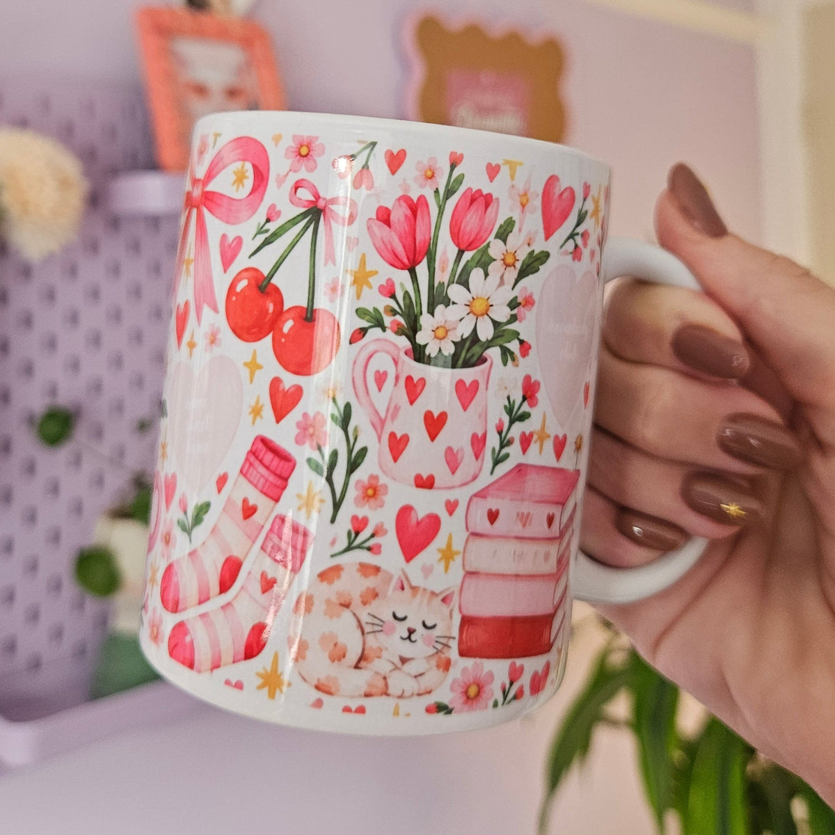 A hand with brown manicured nails holds a white ceramic mug decorated with a pink and red floral, heart, and cherry pattern, set against a pastel purple room background.