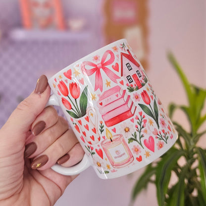 A hand with brown nail polish holds a white ceramic mug decorated with pink tulips, a pink bow, a pink house, and a pink perfume bottle. The background is a soft-focus indoor setting with a lavender wall, a potted green plant, and blurred picture frames.