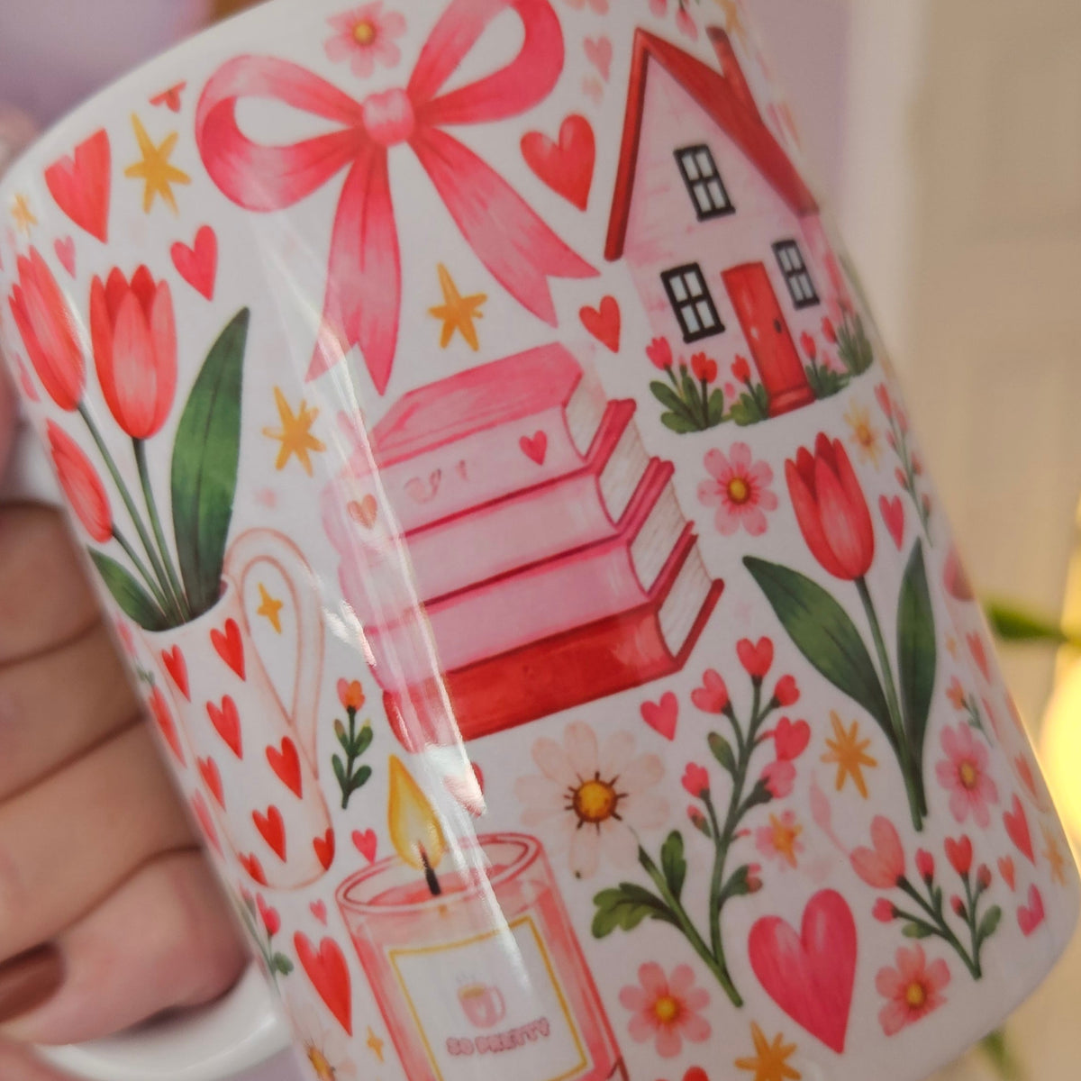 A hand holds a white ceramic mug decorated with a colorful spring-themed illustration featuring pink tulips, red hearts, a pink bow, a small house, and a candle. The background shows a softly blurred indoor setting with a lavender wall and a potted plant.
