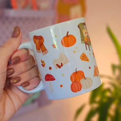  white ceramic mug decorated with autumn illustrations, including pumpkins, a scarf, a book, a mushroom, and a slice of pizza, against a softly blurred indoor background.