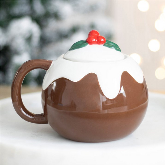 A ceramic mug shaped like a pudding, with a glossy brown body, white icing details, and a small red berry and green holly decoration on top, shown on a light surface with a softly blurred festive background.