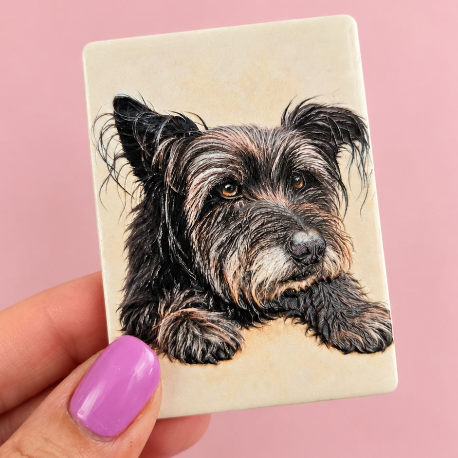 A hand with lavender nail polish holds a square cream-colored ceramic tile featuring a detailed portrait illustration of a black and tan wire-haired dog, set against a solid pink background.