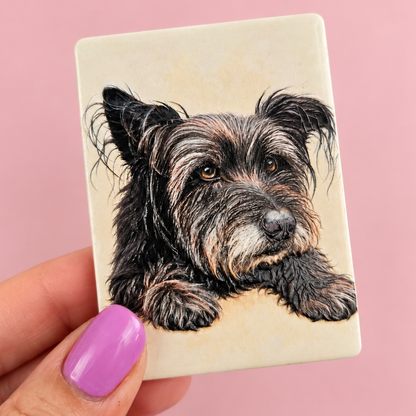 A hand with lavender nail polish holds a square cream-colored ceramic tile featuring a detailed portrait illustration of a black and tan wire-haired dog, set against a solid pink background.