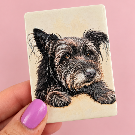 A hand with lavender nail polish holds a square cream-colored ceramic tile featuring a detailed portrait illustration of a black and tan wire-haired dog, set against a solid pink background.