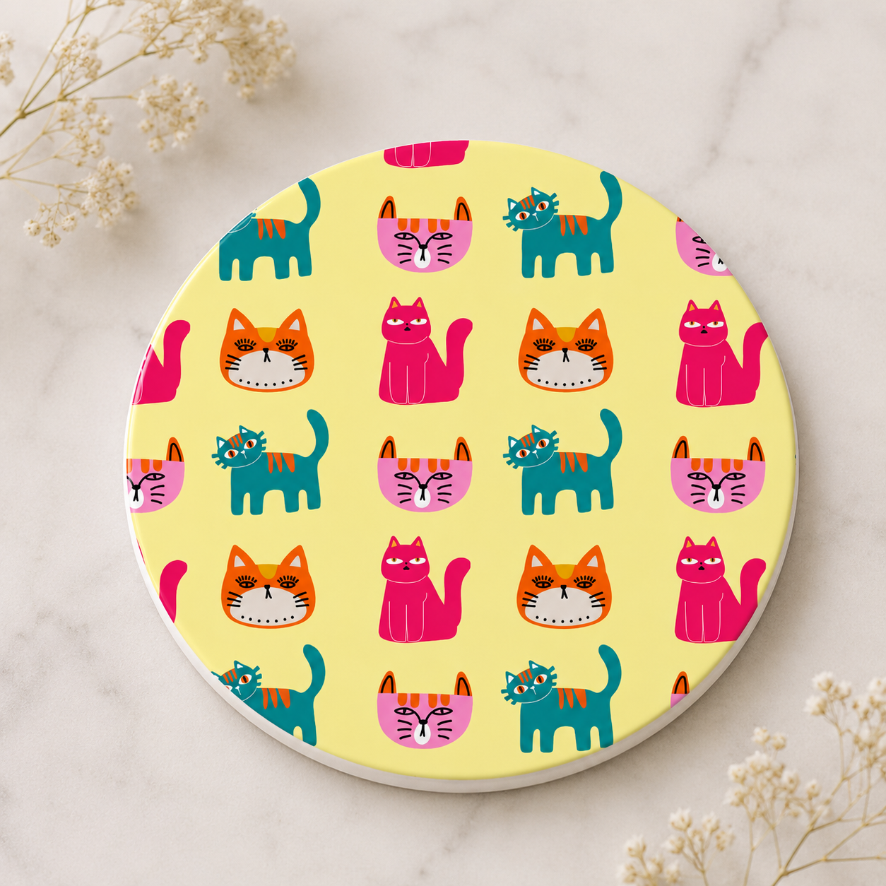 A round coaster with a playful cat pattern on a pale yellow background, styled on a light marble surface with small dried flowers in the corners.