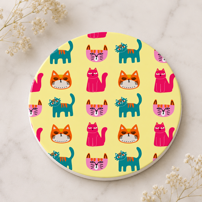 A round coaster with a playful cat pattern on a pale yellow background, styled on a light marble surface with small dried flowers in the corners.