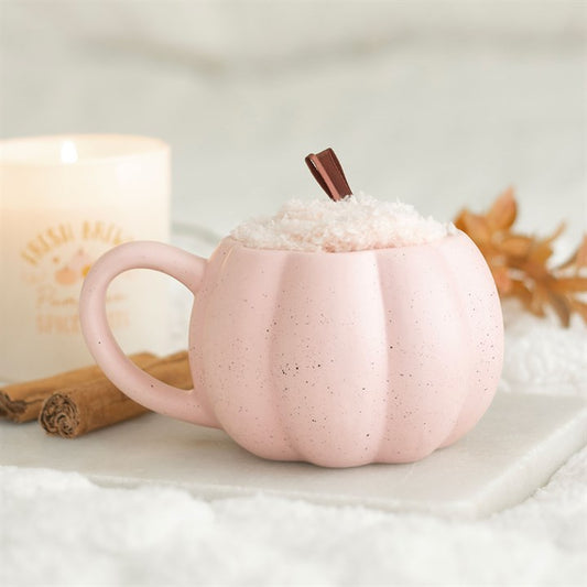 Pink pumpkin-shaped mug with a cinnamon stick on a white surface with a blurred candle in the background.