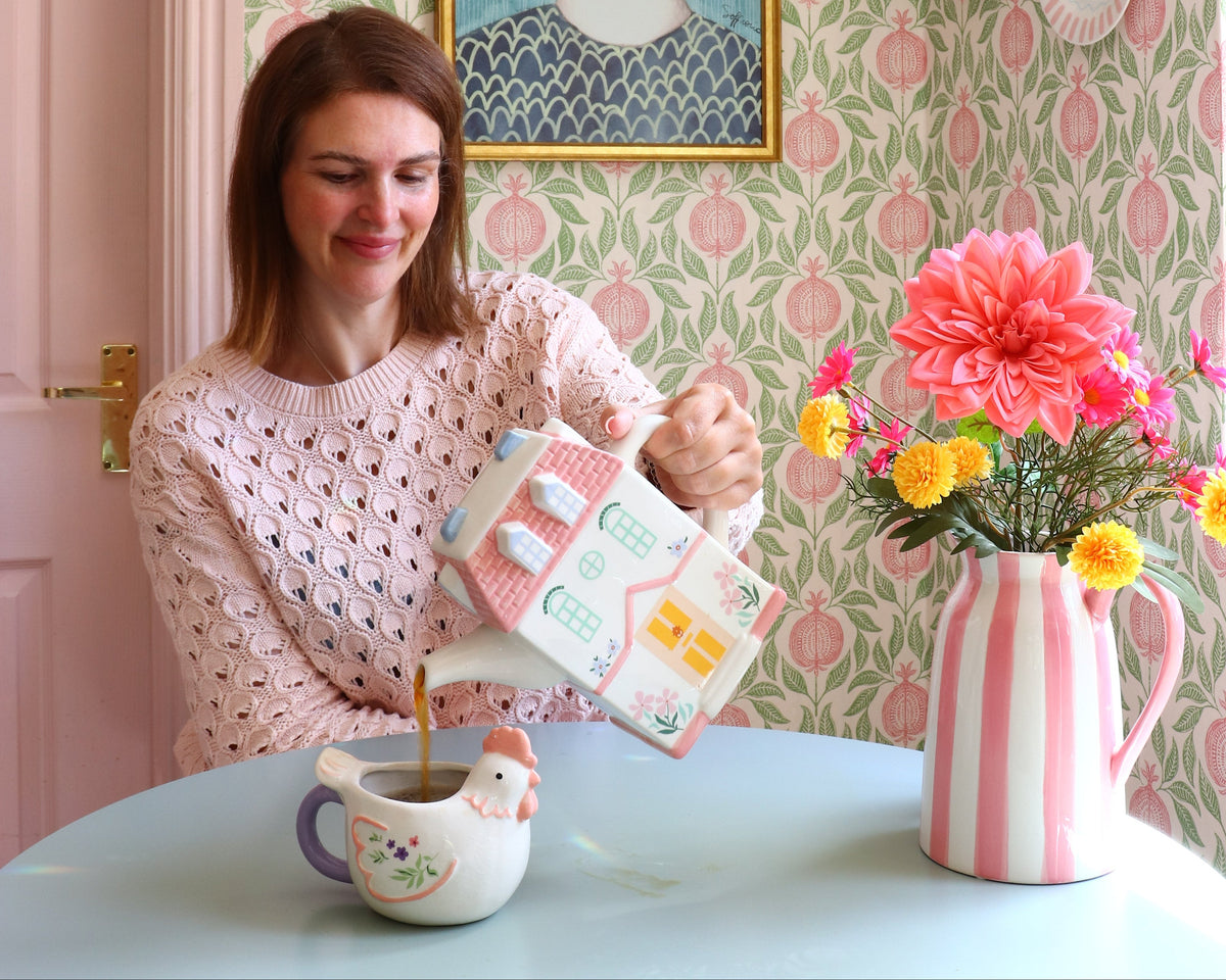 A woman in a pink patterned top sits at a light blue table holding a pink striped mug and a small box, with a floral wallpapered wall, framed portrait, and decorative plates in the background.