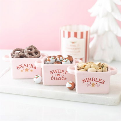 Three pink ceramic bowls labeled 'Snacks', 'Sweet treats', and 'Nibbles' filled with various snacks on a white surface.