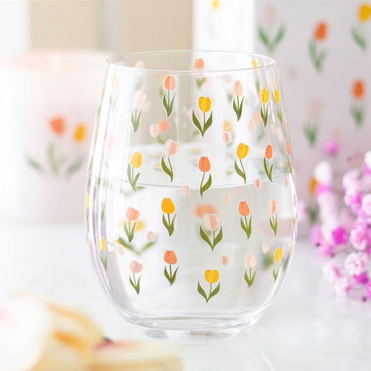 A clear stemless wine glass decorated with a repeating pattern of small tulips in orange, pink, yellow, and green, set against a softly blurred background with flowers.