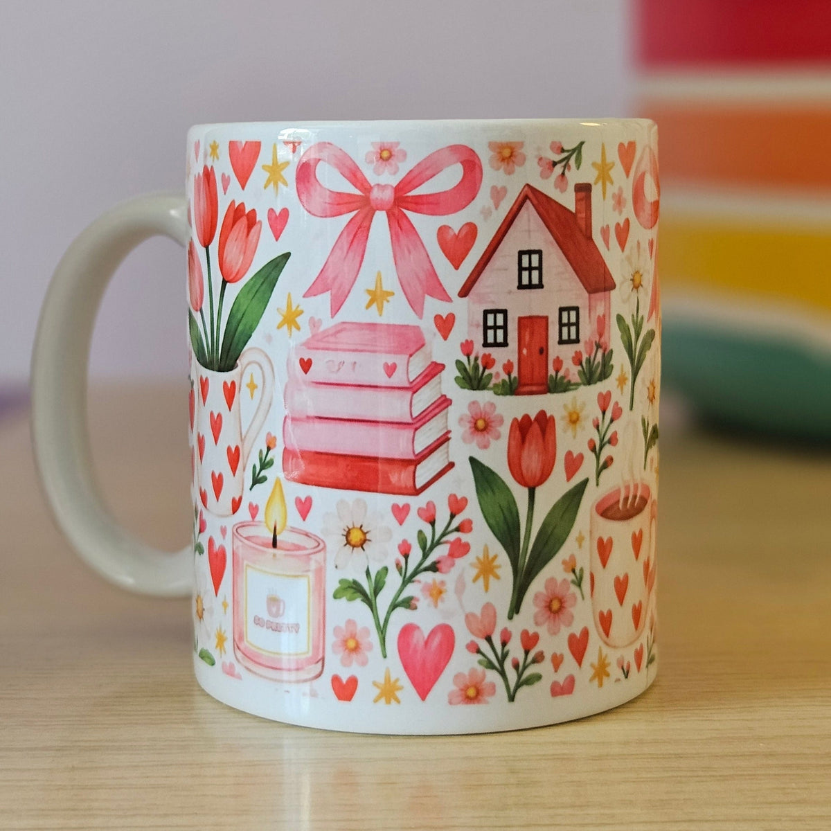 A white ceramic mug decorated with a whimsical illustration of a pink gift box, a small house, tulips, hearts, and stars, sitting on a light wooden table.