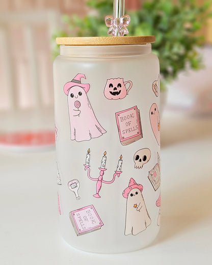 A frosted glass tumbler with a bamboo lid and a clear glass straw, decorated with cute pink and white Halloween illustrations including ghosts, a pumpkin, a witch hat, and candles. The tumbler is set on a white surface with a softly blurred background of a plant and curtains.